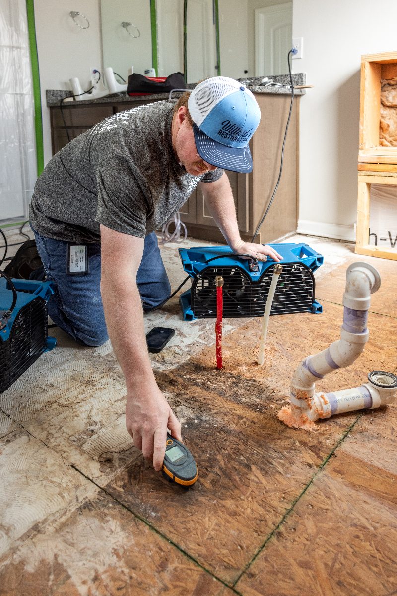 A technician taking a moisture reading with air movers running on an exposed subfloor